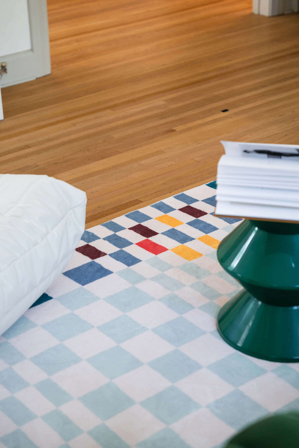 View of the multicolor checkered rug corner under a green pedestal table, with warm wood flooring and cozy room setting.