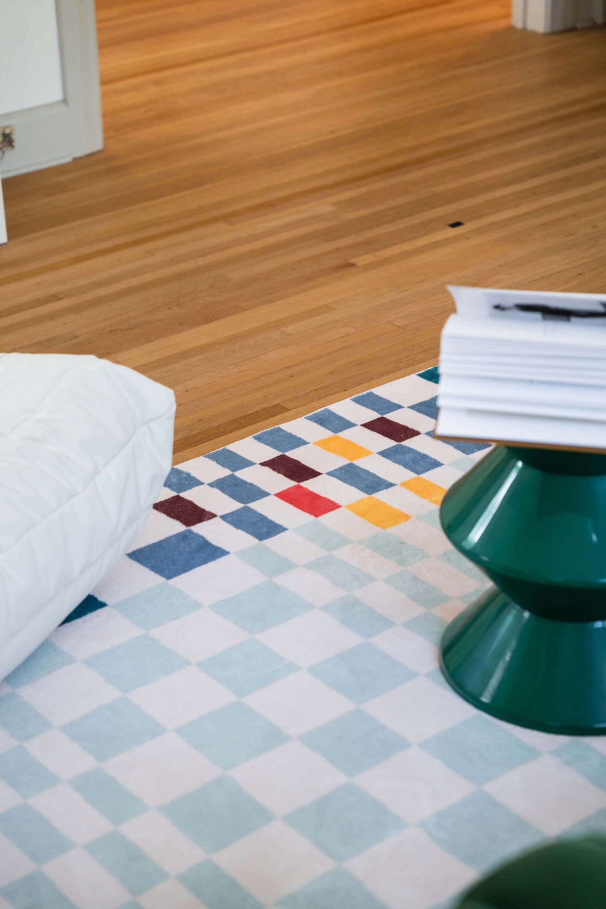 View of the multicolor checkered rug corner under a green pedestal table, with warm wood flooring and cozy room setting.