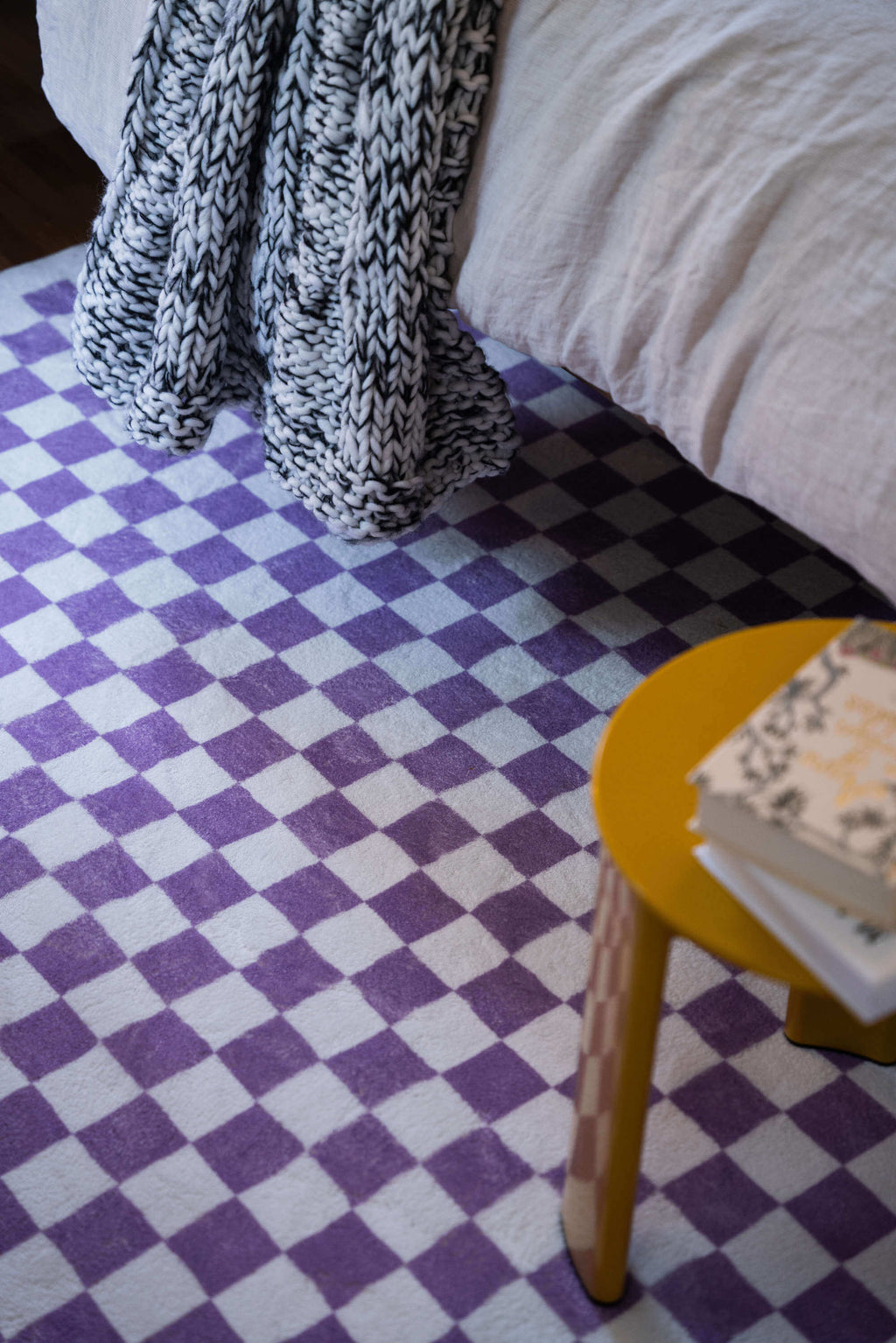 Purple checkered rug under a cozy bed and chunky knit throw, creating a warm, textural corner with pops of color and comfort.