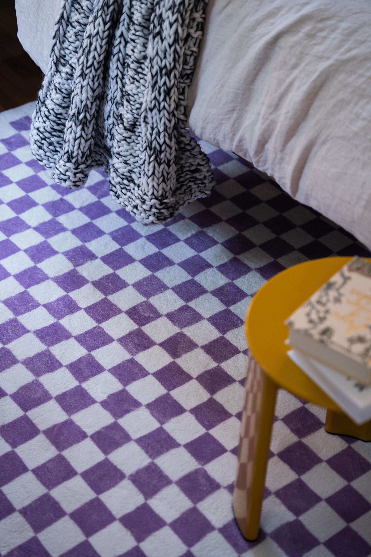 Purple checkered rug under a cozy bed and chunky knit throw, creating a warm, textural corner with pops of color and comfort.