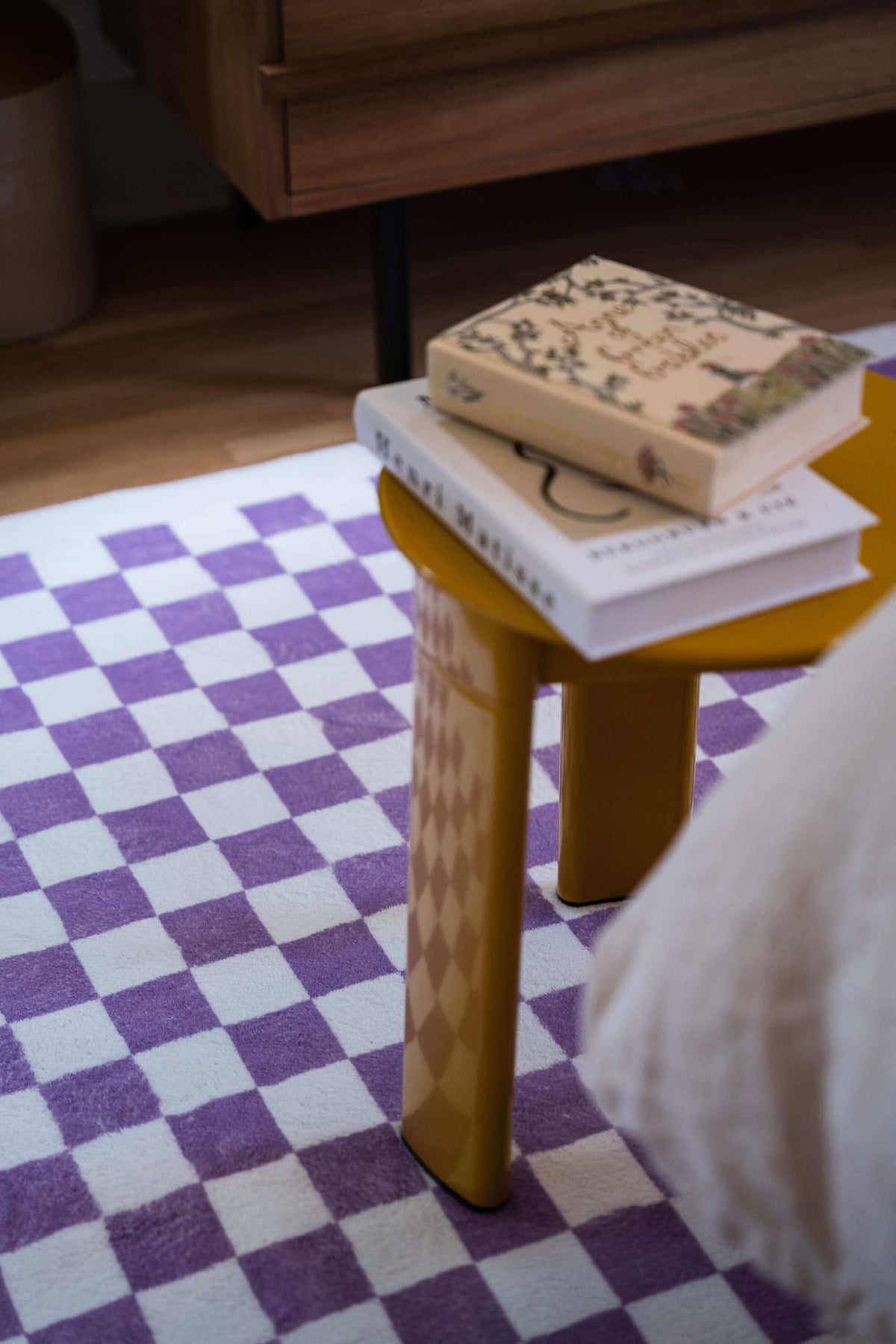 Close-up of a purple checkered rug next to a mustard yellow side table with stacked books, adding playful contrast to the room.