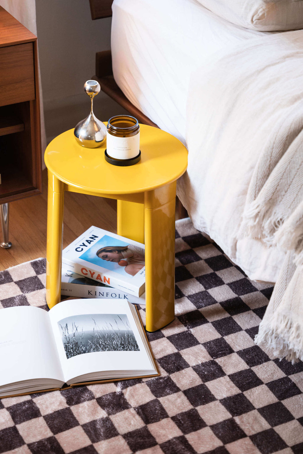A cozy bedroom corner styled with the Chocolate Brown Checkered Rug, yellow side table, books, and soft layered bedding.