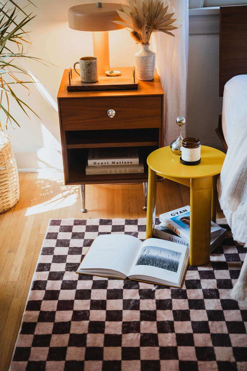 Close-up of the checkered rug used as a bedside mat, styled with a yellow table, books, and warm lighting.