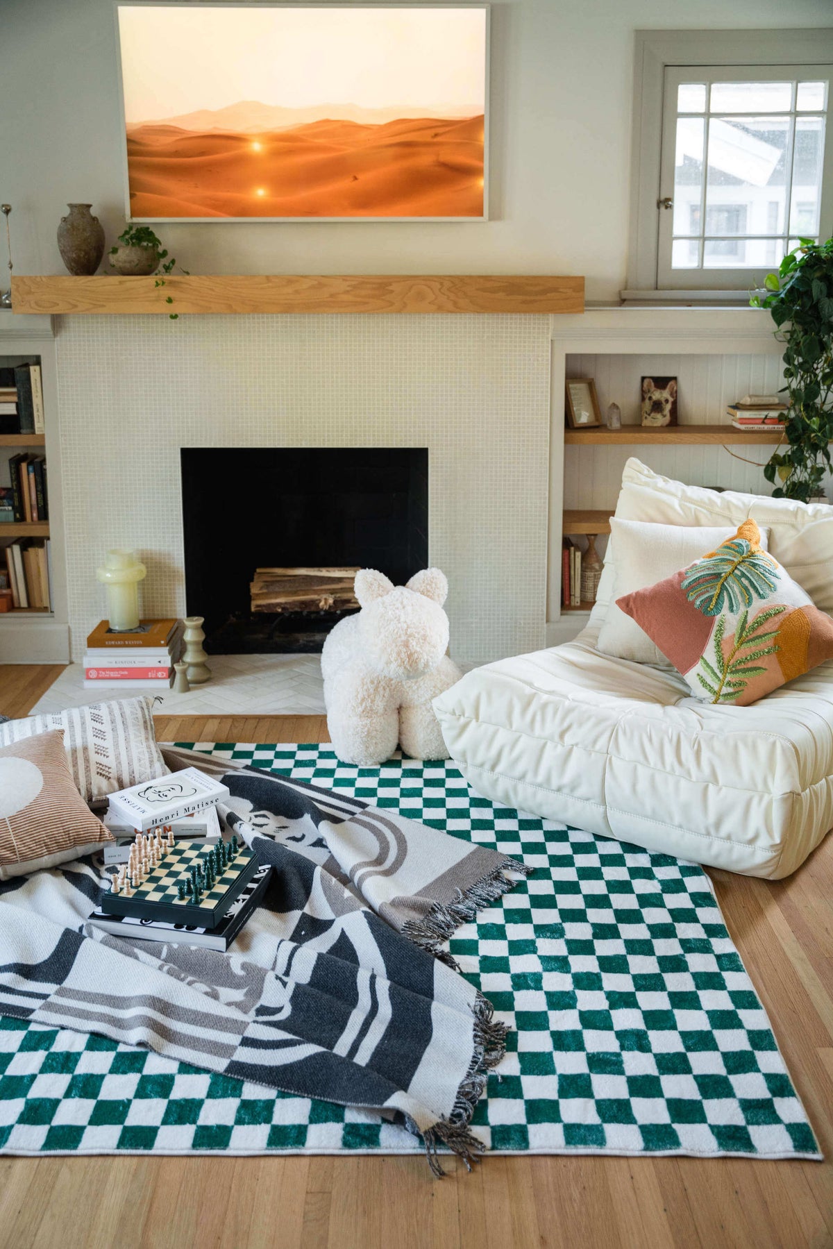 Checkered rug placed by the fireplace, styled with a white lounge chair, throw blanket, and a chess set on the floor.