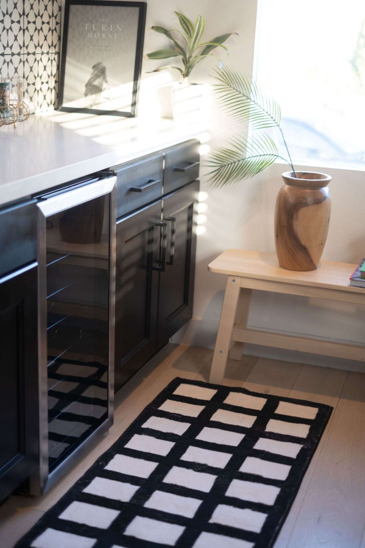 Black and white grid runner under soft sunlight, next to black lower cabinets and a decorative wood bench.