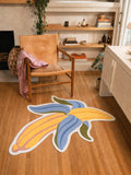 4' × 5' half-peeled banana rug with yellow fruit, blue peel, green tips, and a white border beside a wooden chair.