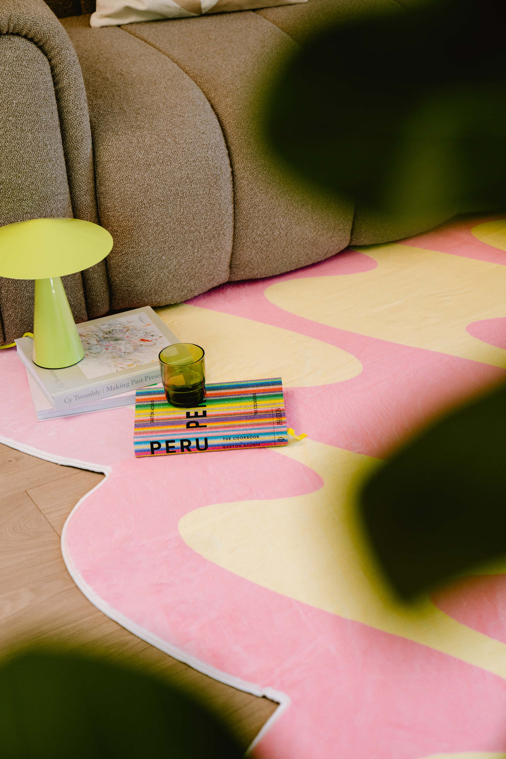 A stack of books and a green glass sit beside a lime green table lamp on a pink and yellow wavy rug near a brown sofa.