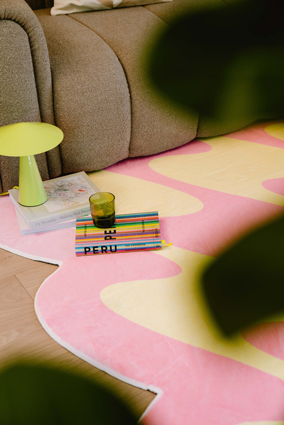 A stack of books and a green glass sit beside a lime green table lamp on a pink and yellow wavy rug near a brown sofa.