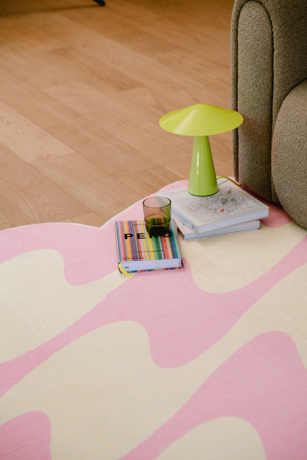 Corner of pink and yellow wavy rug with green mushroom lamp, books and glass beside sofa on wood floor.