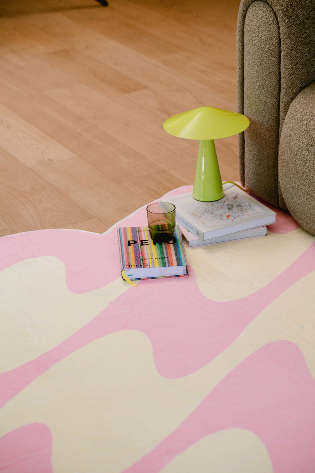 Corner of pink and yellow wavy rug with green mushroom lamp, books and glass beside sofa on wood floor.