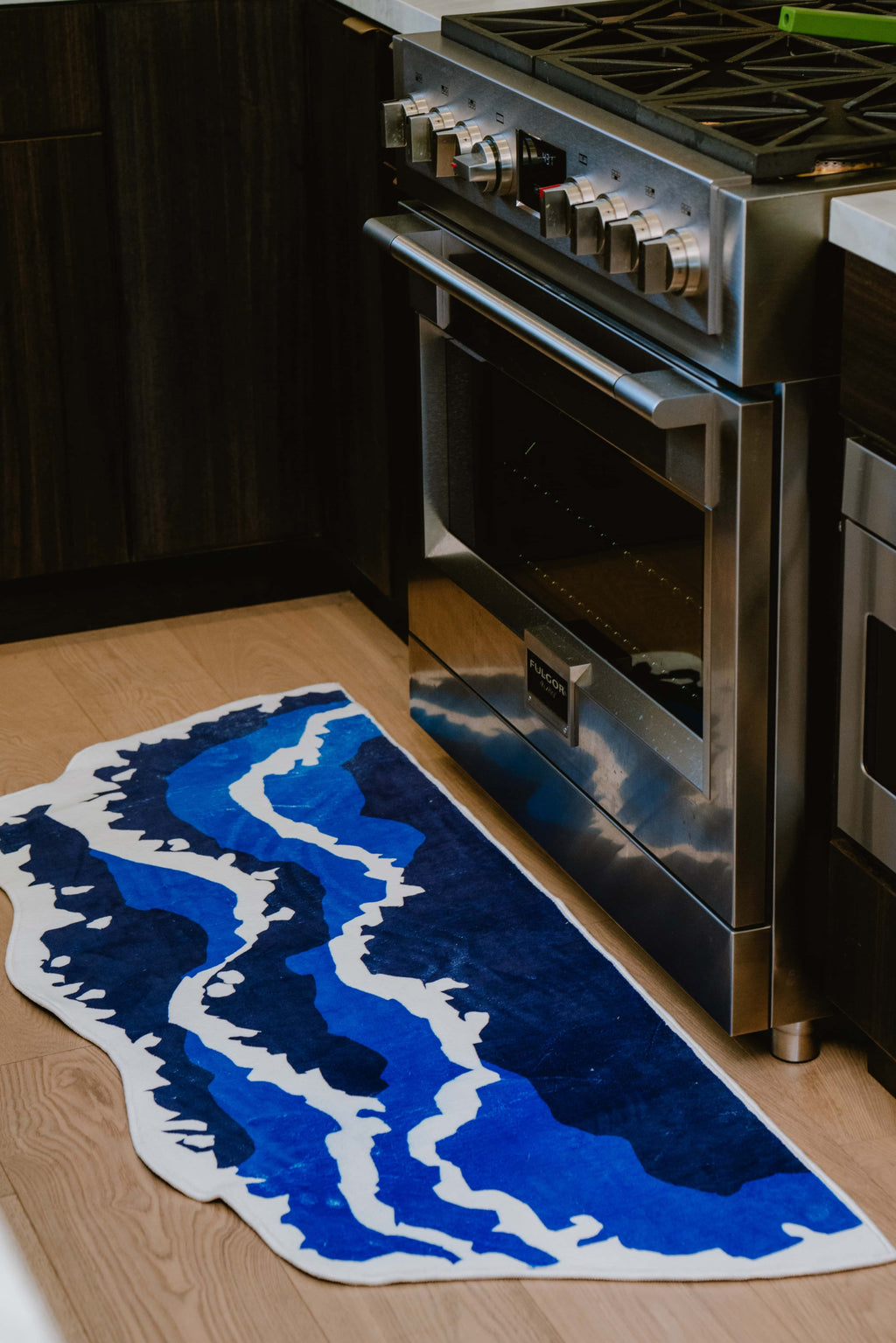 Closer look at the irregular blue wavy runner rug laid near a stainless steel oven on a light-toned kitchen floor.