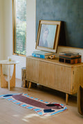 Brown dog runner rug next to a wood media console with framed art and books, in a bright sunlit corner.