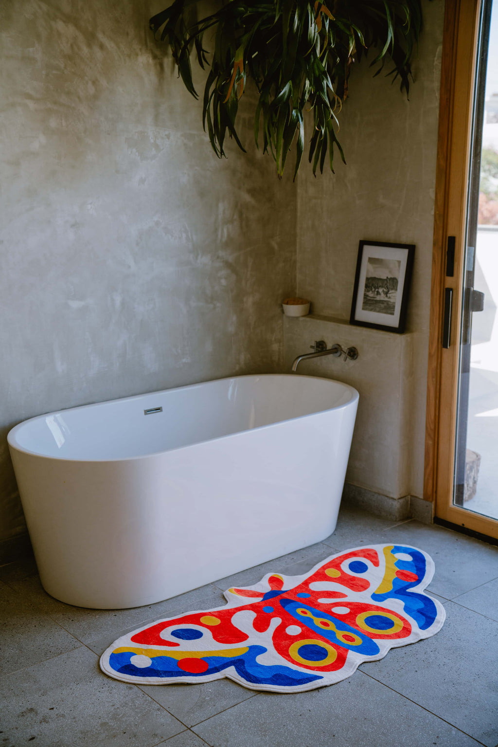 Colorful butterfly rug beside a soaking tub on stone tiles, tucked in a corner with textured walls and natural light.
