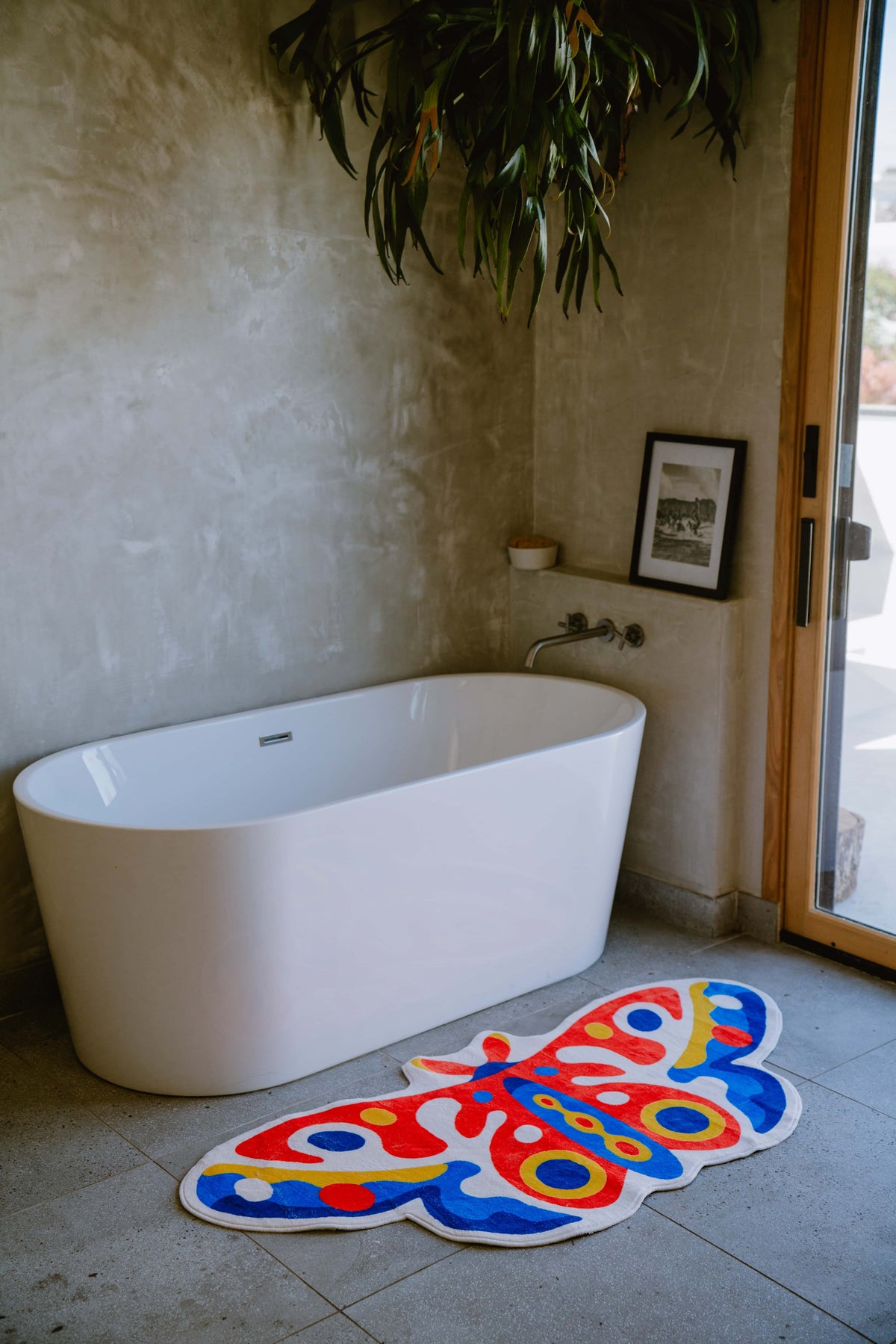 Colorful butterfly rug beside a soaking tub on stone tiles, tucked in a corner with textured walls and natural light.