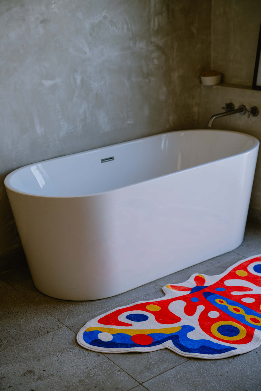 Butterfly rug on gray tiles beside a freestanding tub, in a textured corner with a soft industrial finish.