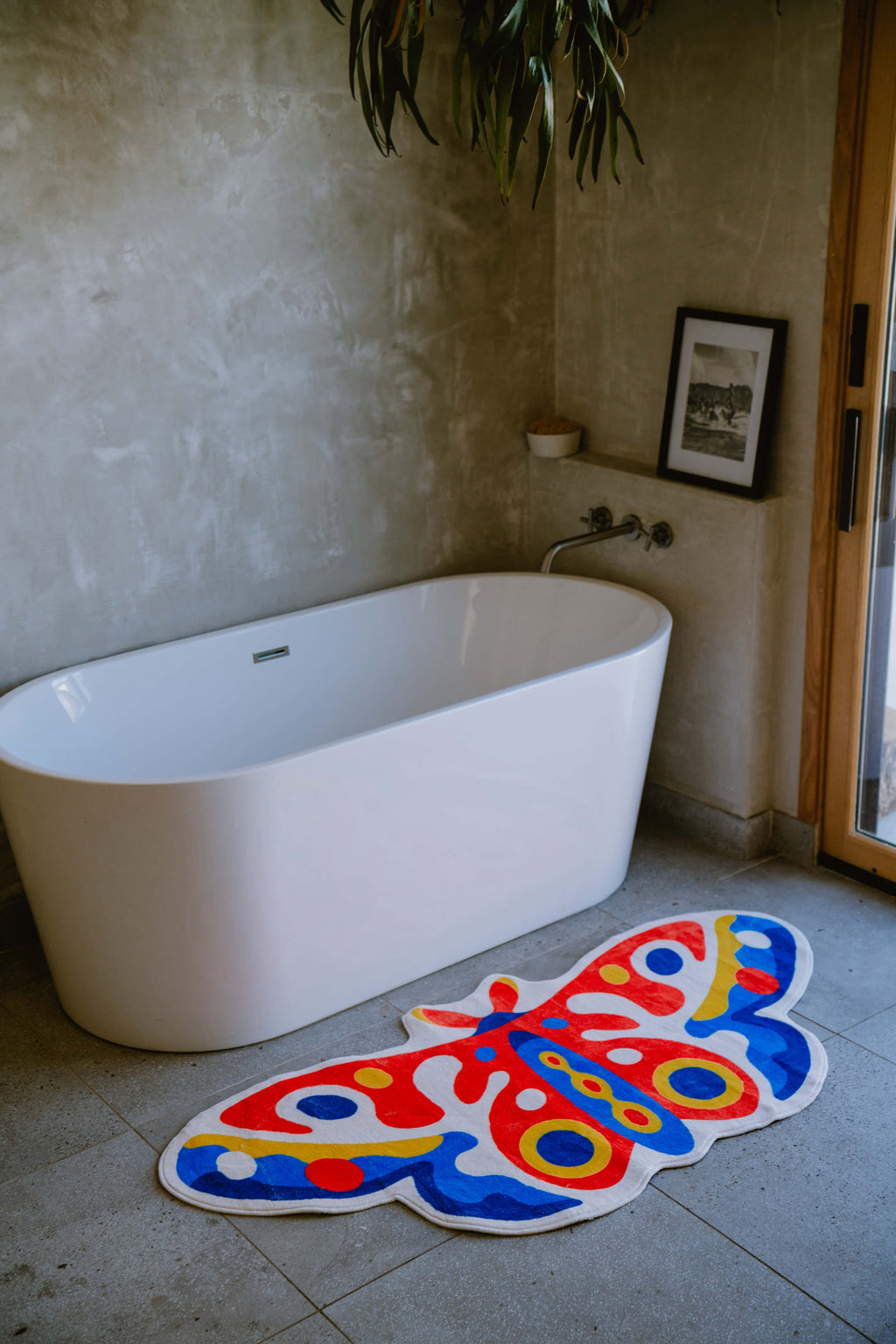 Irregular Butterfly Rug by Johann Banta placed on stone floor beside a freestanding tub in a textured corner space.