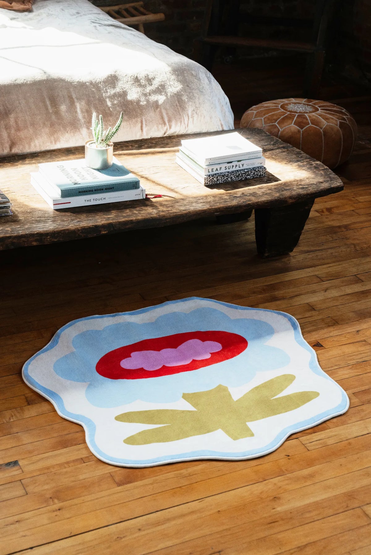 Playful flower rug in blue tones styled near stacked books and pouf, catching sunlight on polished floorboards.