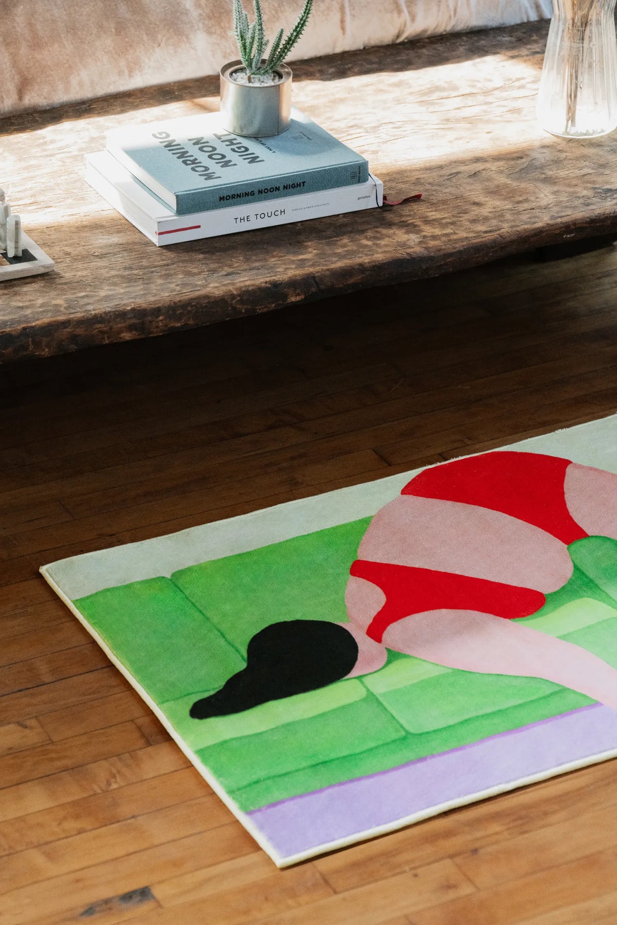Abstract body art rug in vivid red and green tones, styled next to a rustic wooden bench with books and a glass vase.