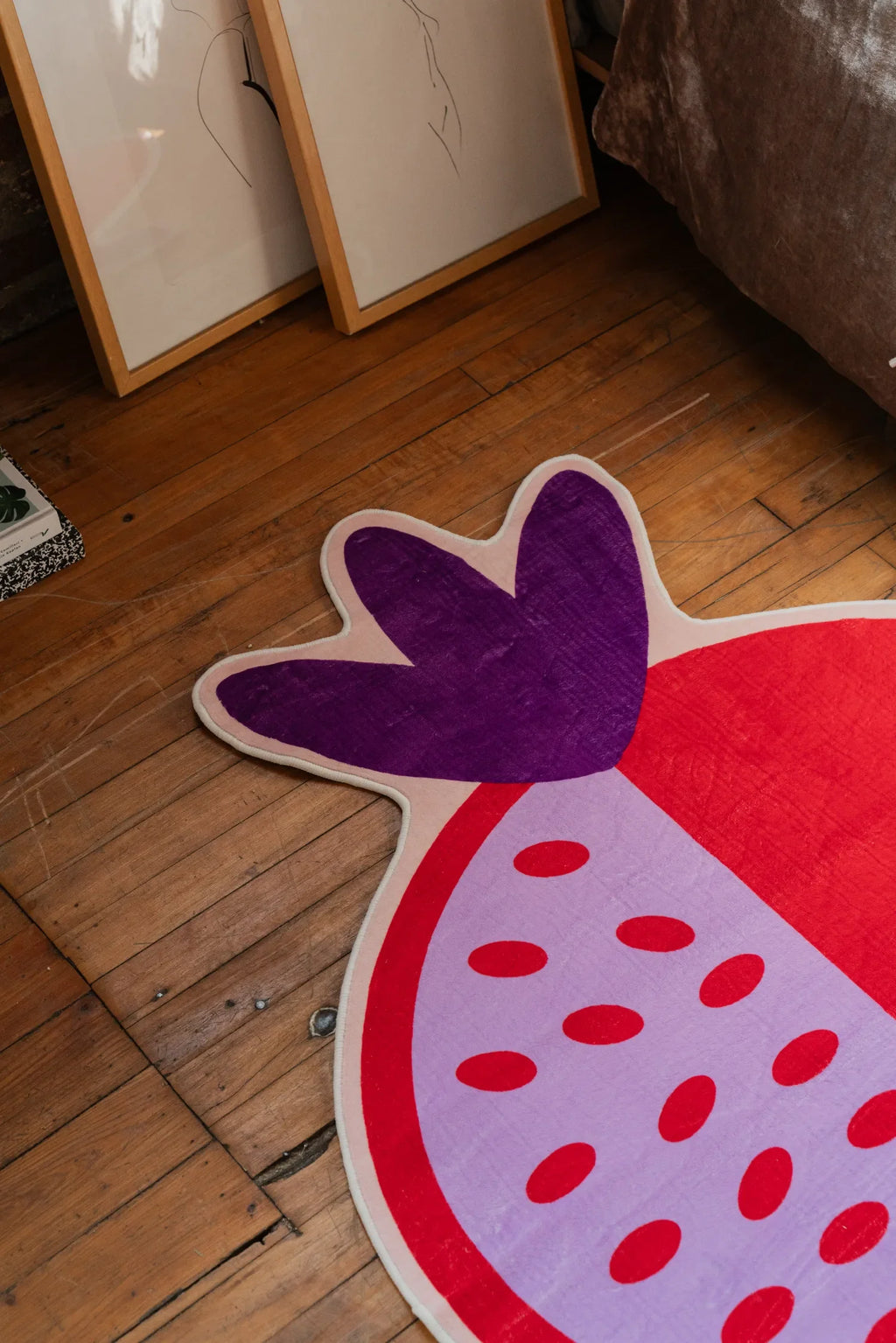 Close-up of the Red Pomegranate Shaped Irregular Rug by Shayla Goethe, placed on a wooden floor near leaning framed artwork.