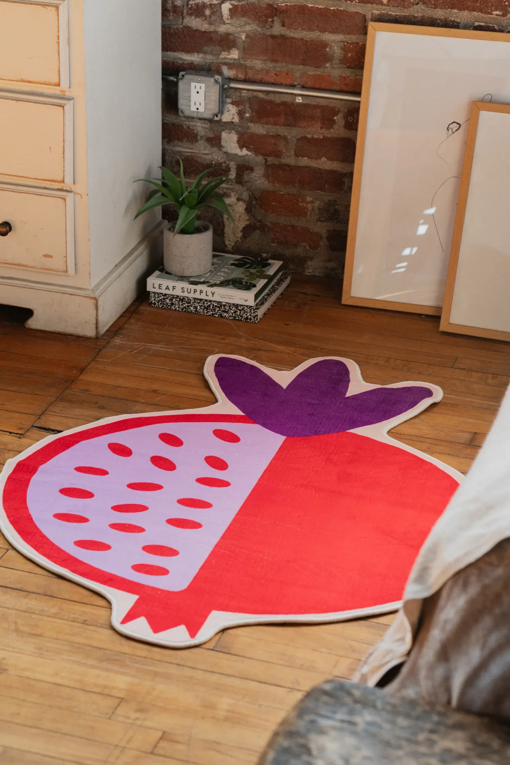 Red pomegranate rug with purple crown detail, styled beside a brick wall and white cabinet in a cozy bedroom corner.