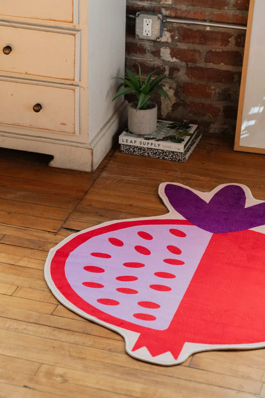 Playful pomegranate-shaped rug with bold red and lavender details, lying on hardwood floor next to a small plant and framed art.