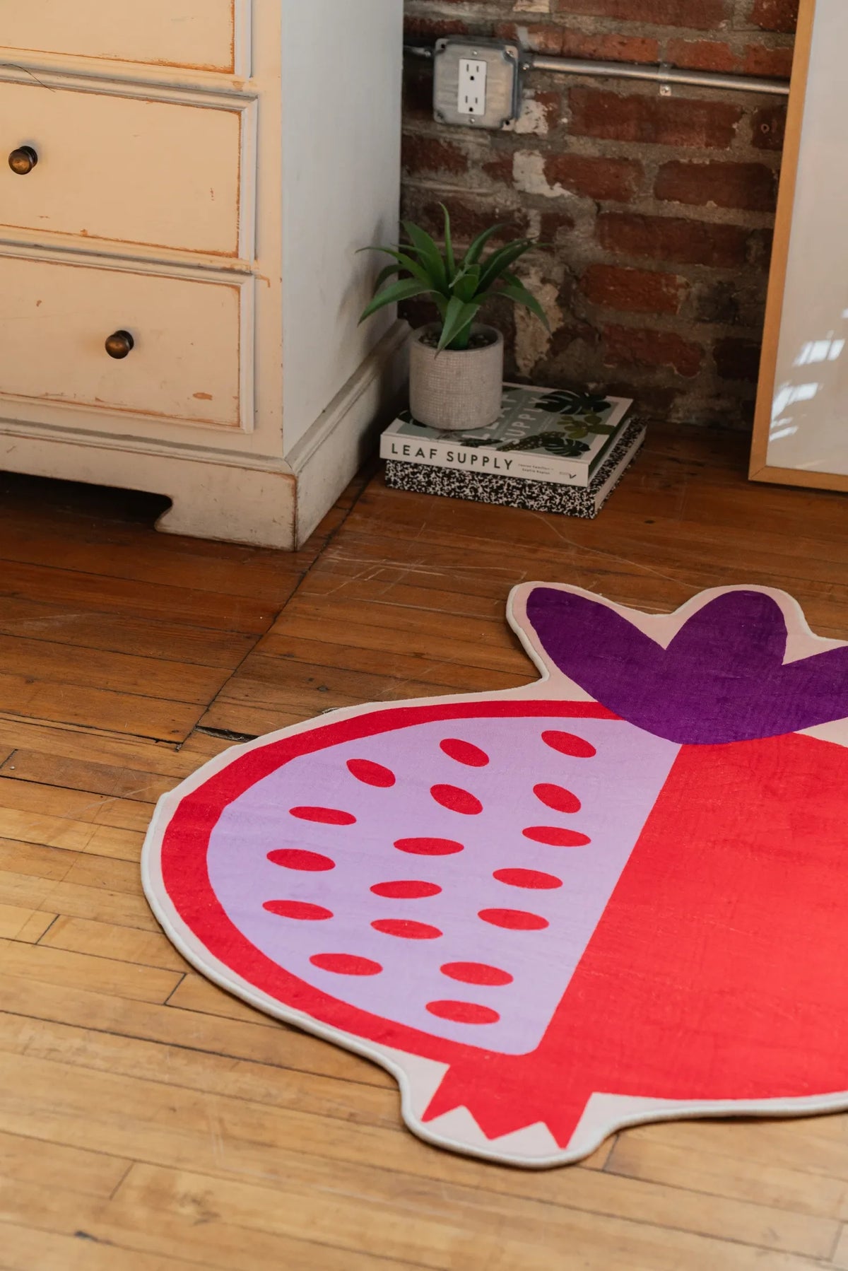 Playful pomegranate-shaped rug with bold red and lavender details, lying on hardwood floor next to a small plant and framed art.