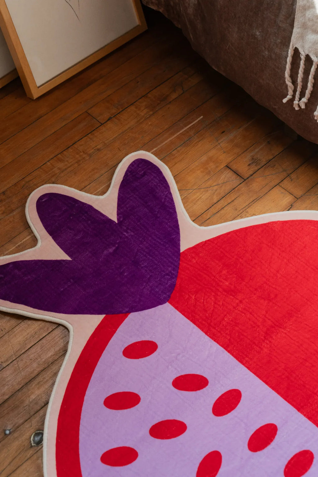 Close-up of pomegranate shaped rug showing purple crown detail beside bed on wooden floor.