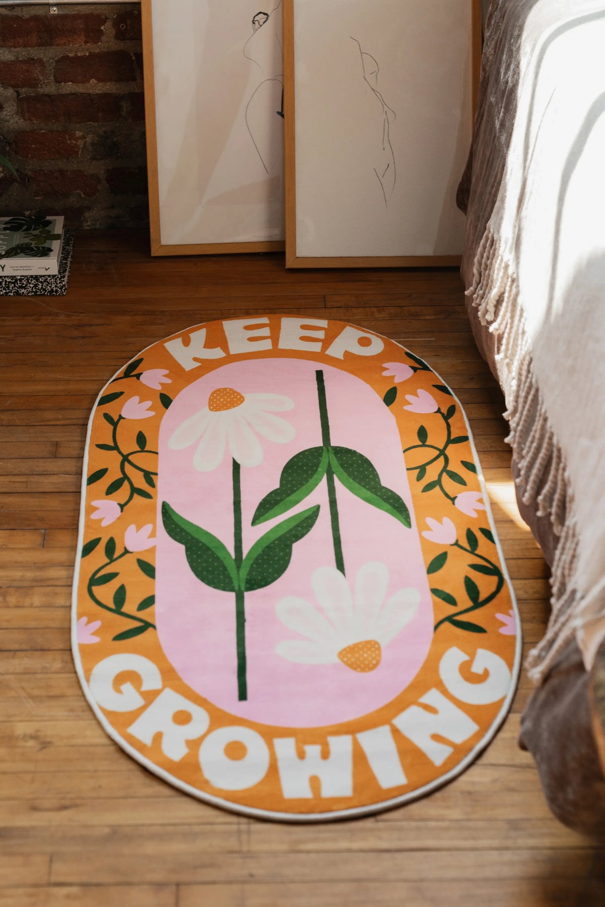 Oval funky cat rug style with floral “Keep Growing” print lying on wooden floor by artwork frames in a sunlit corner