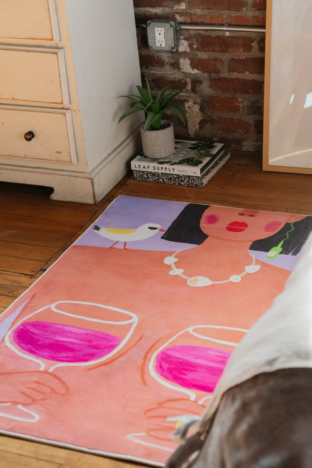A funky art rug with bold wine and woman design set near potted plant and stacked books against a brick wall.