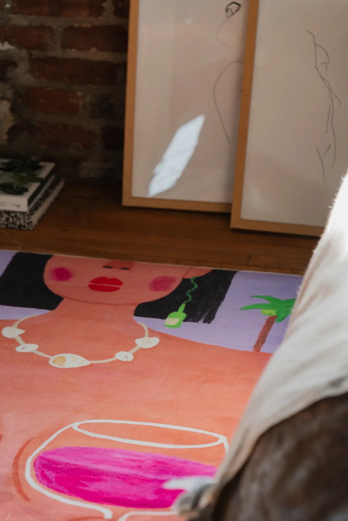 Playful rug showing woman with necklace and wine glasses placed by leaning sketches and warm sunlight on wood floor.