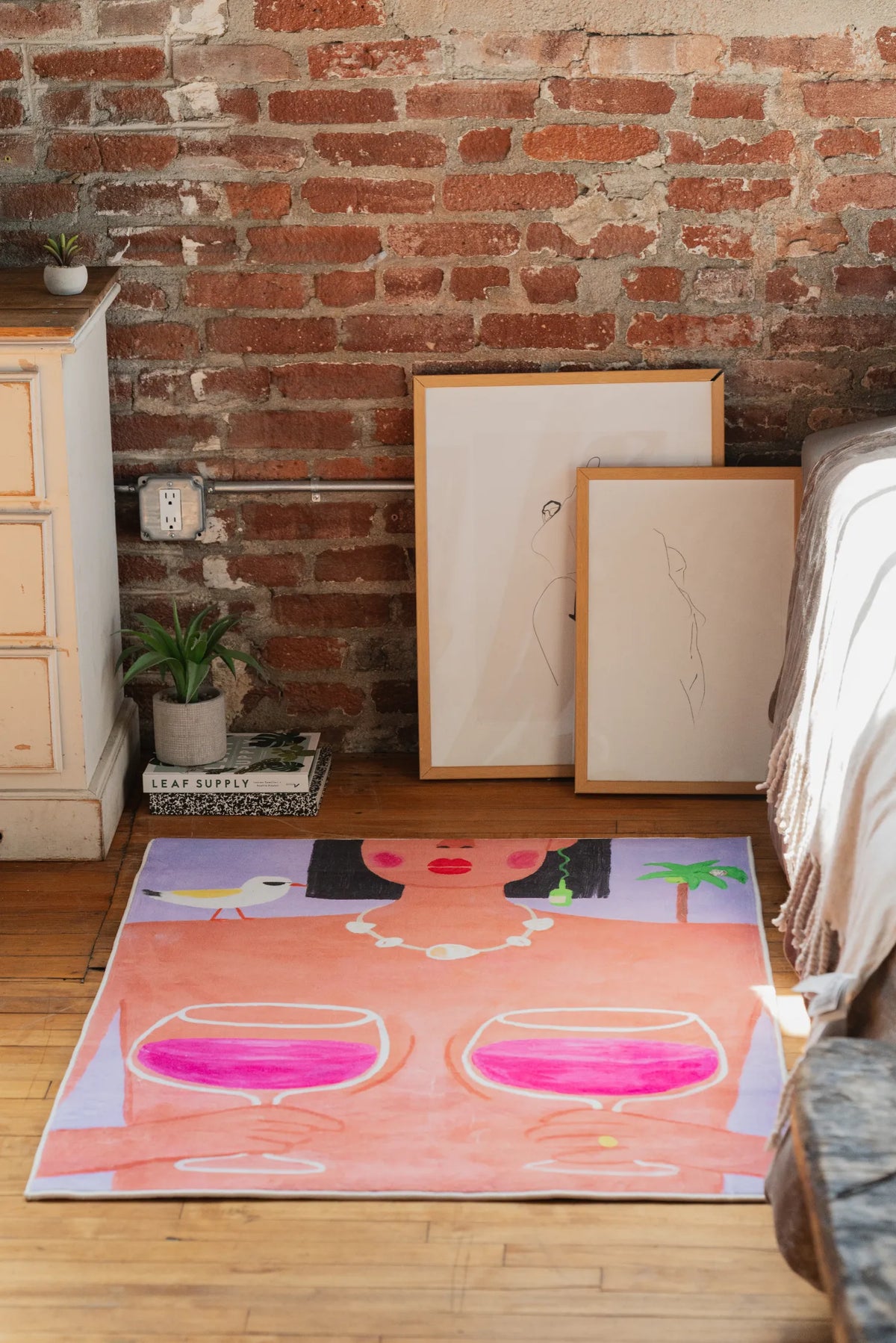 Funky Wine and Woman Art Rug by Nimei resting under framed art and brick wall beside a soft bed corner.