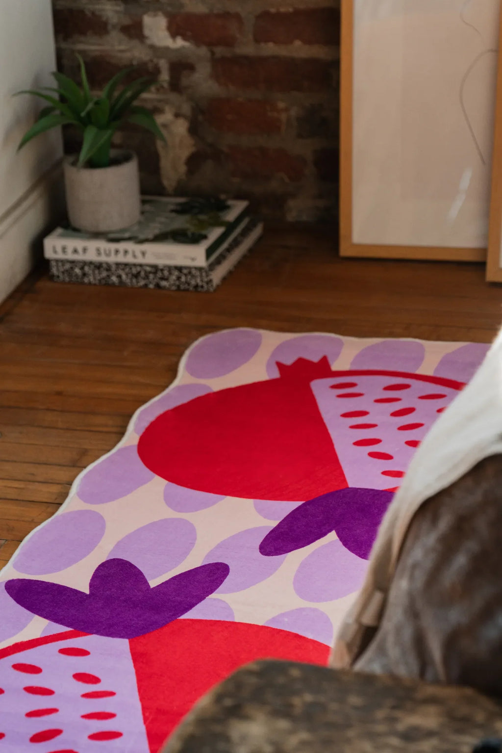 Playful pomegranate fruit rug in red and purple tones, laid near a potted plant and framed artwork on wooden floor.