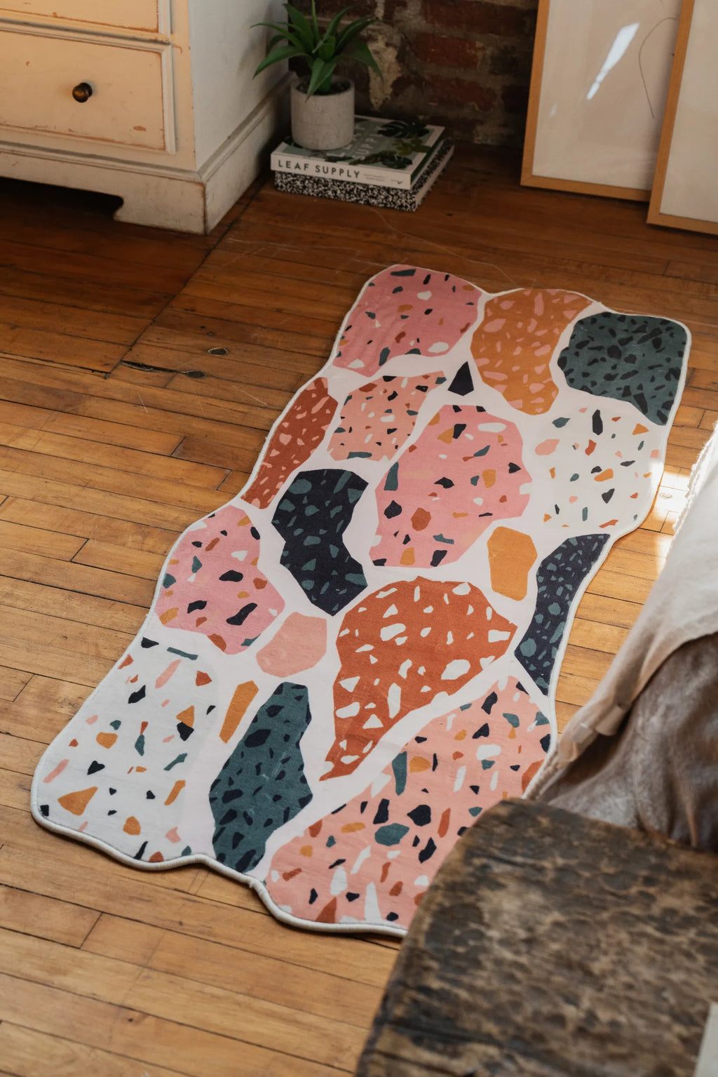 A mosaic abstract runner rug is placed on the wood floor beside a bed and a dresser against a brick wall.