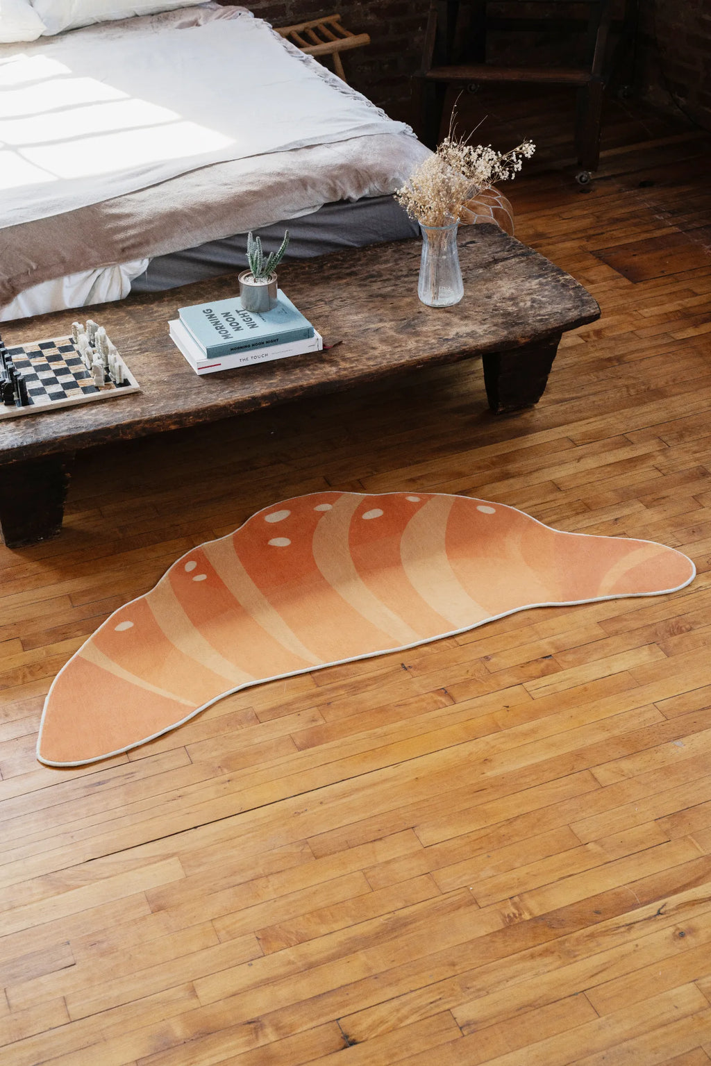 Buttery Orange Croissant Rug by Petra Lizde on a wooden floor near a rustic bench, with books and a vase in the background