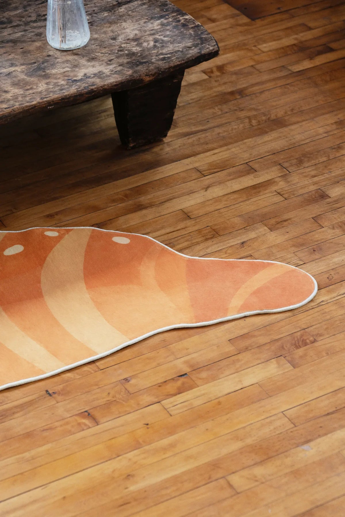 Close-up of the croissant rug on wooden floor, highlighting its orange gradient and white dotted details beside a rustic table