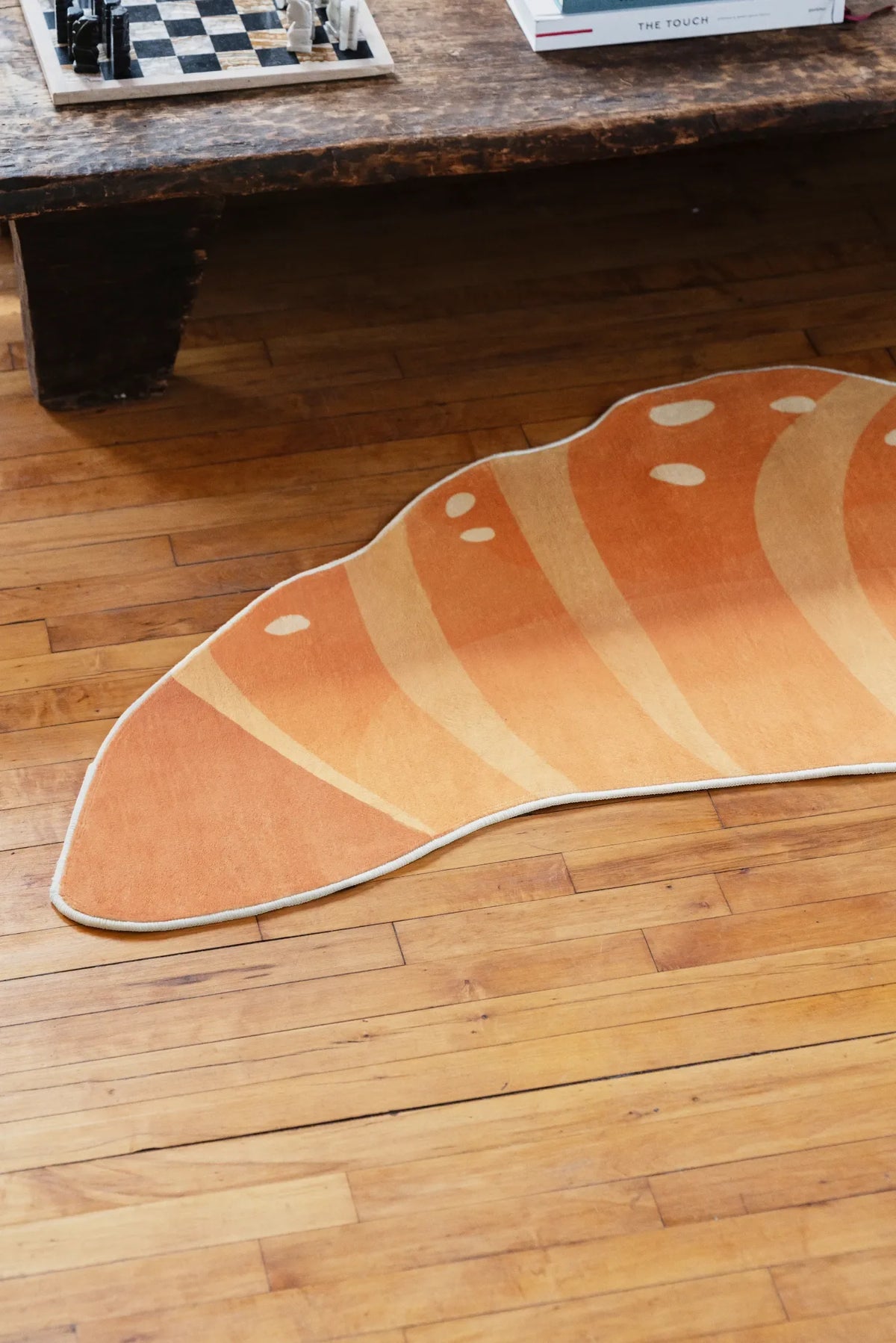 Croissant-shaped rug partially near a rustic wooden bench on a wood floor, with a chessboard and decor items on the bench above