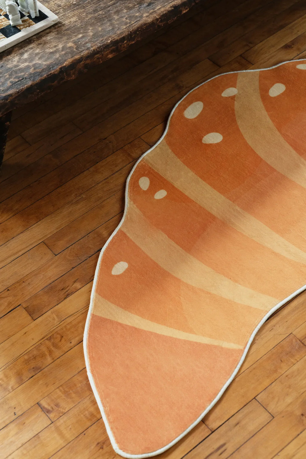 Croissant rug shown close-up on a wooden floor, with its orange gradient and dotted pattern partly framed by a rustic bench