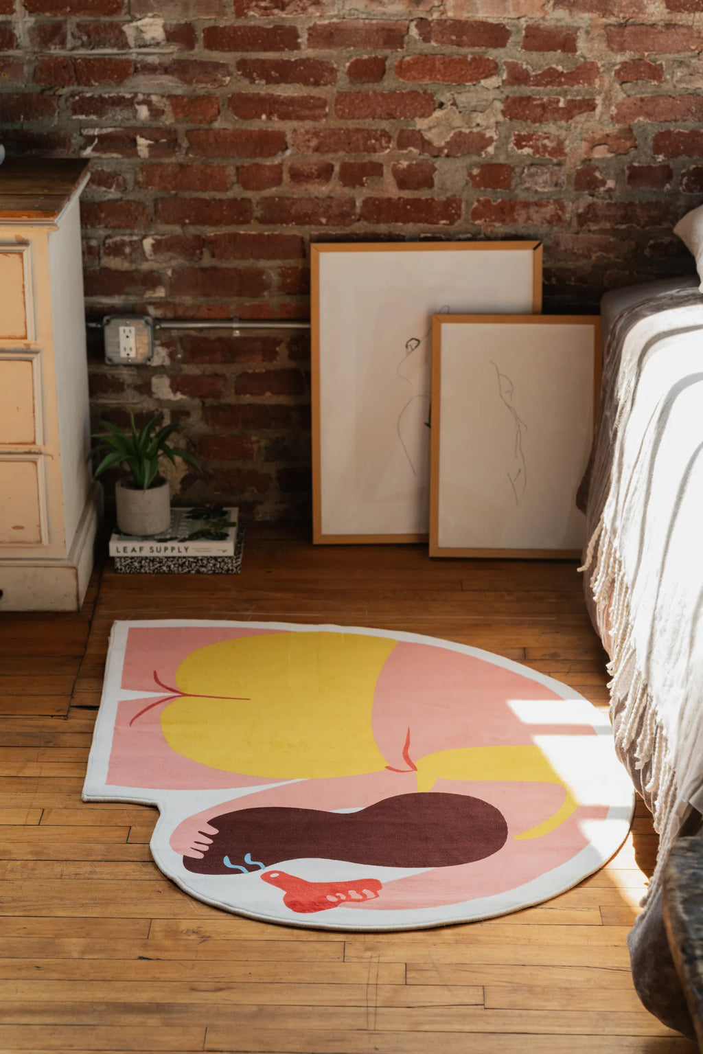 Round body art rug beside bed and brick wall, shot diagonally to show bold yellow and pink design.