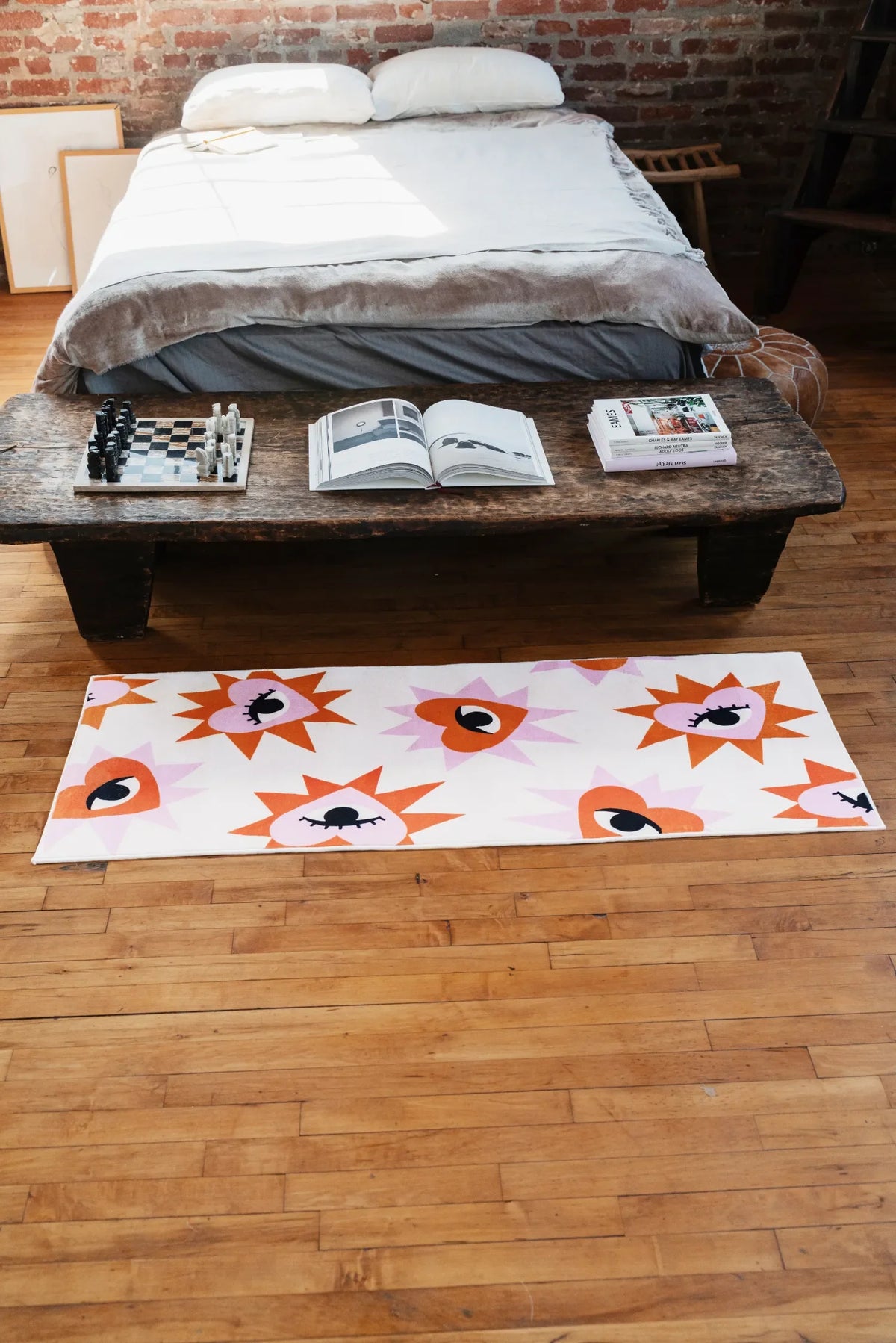 Heart eye rug styled at the foot of rustic bed with wood table and open book and chess set creating a cozy bedroom look