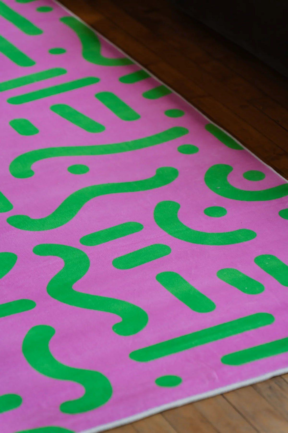 Retro graphic rug with pink and green pattern placed against wood floor near a dark-toned sofa.