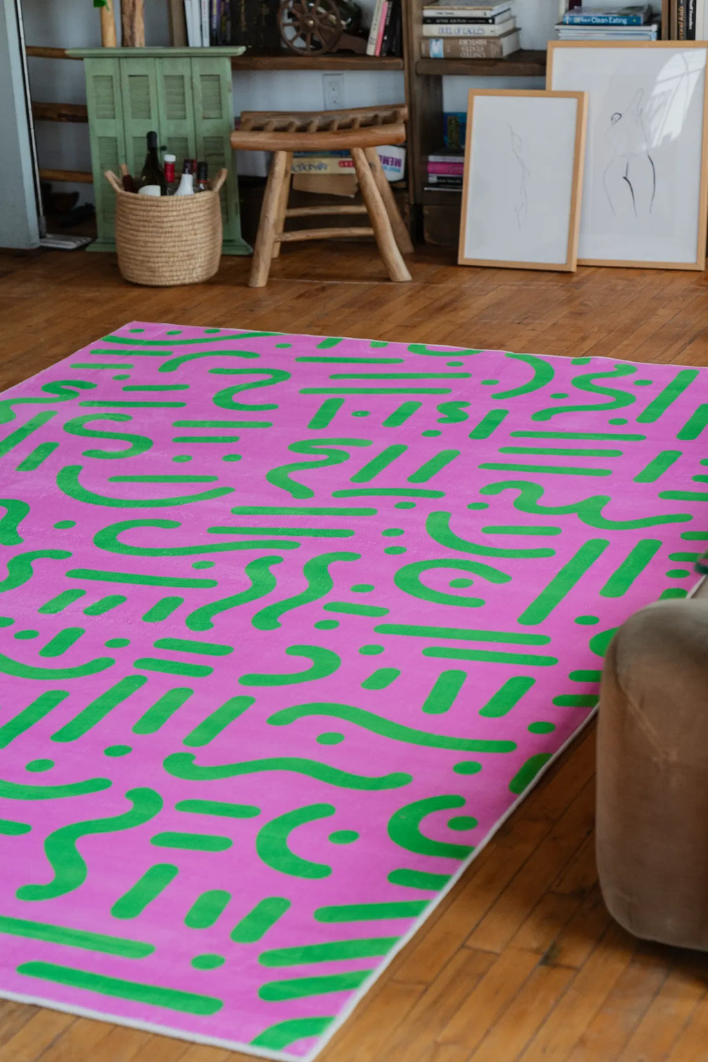 Pink and green retro graphic area rug adds a bold pop of color beside a wooden stool and framed sketches on wood floor.