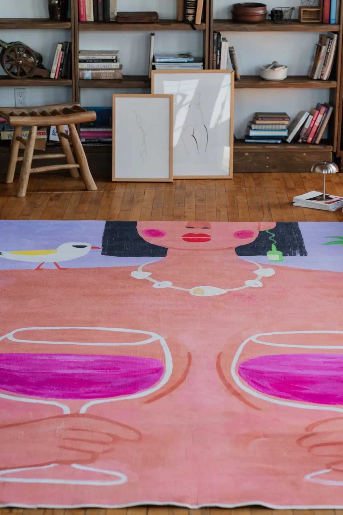 Funky Wine and Woman Art Rug by Nimei set on wood floor in front of tall bookshelf with framed sketches leaning below.