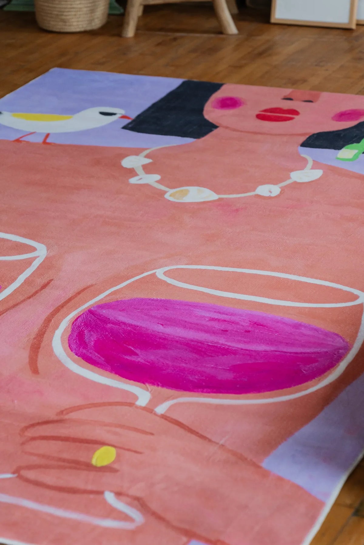 Playful rug featuring bold wine glasses and woman portrait viewed at angle near basket and stacked art books on floor.