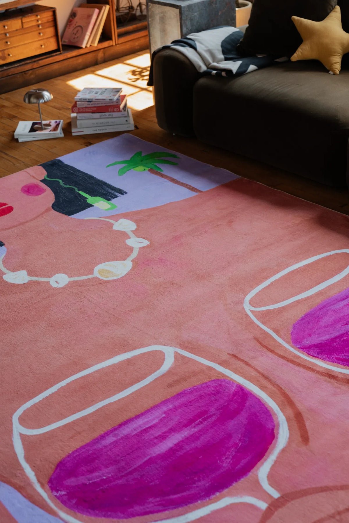 Funky Wine and Woman Art Rug spread across wood floor near dark sofa with star pillow and stacked books.
