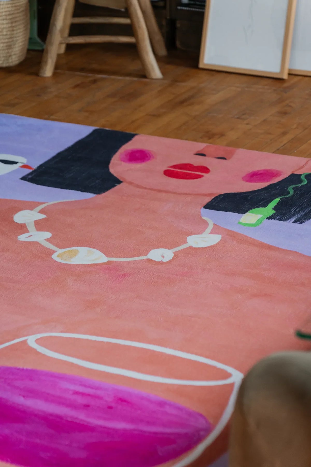 A funky art rug showing bold woman portrait and wine motif captured from low angle beside rustic stool and sketches.