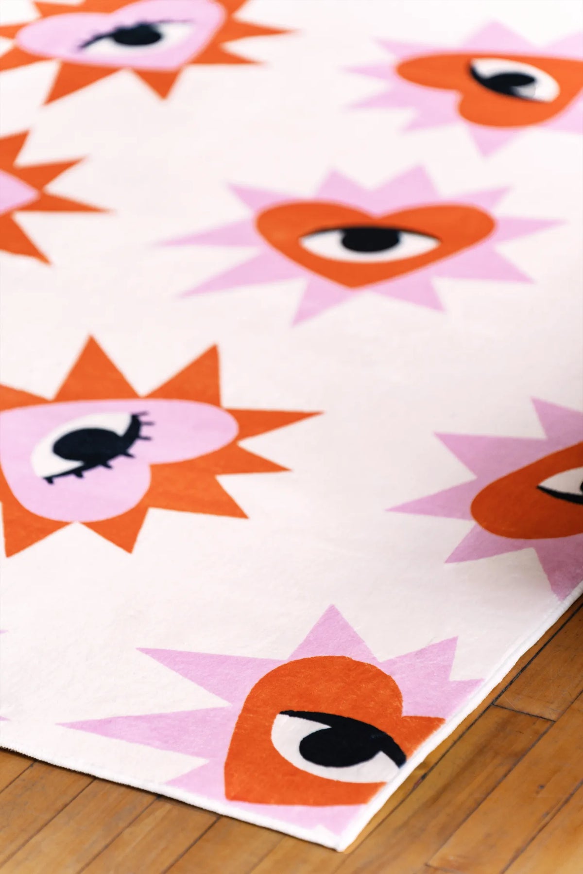 Close-up view of modern area rug showing bold red and pink heart eye motifs resting on warm wooden floor with soft texture