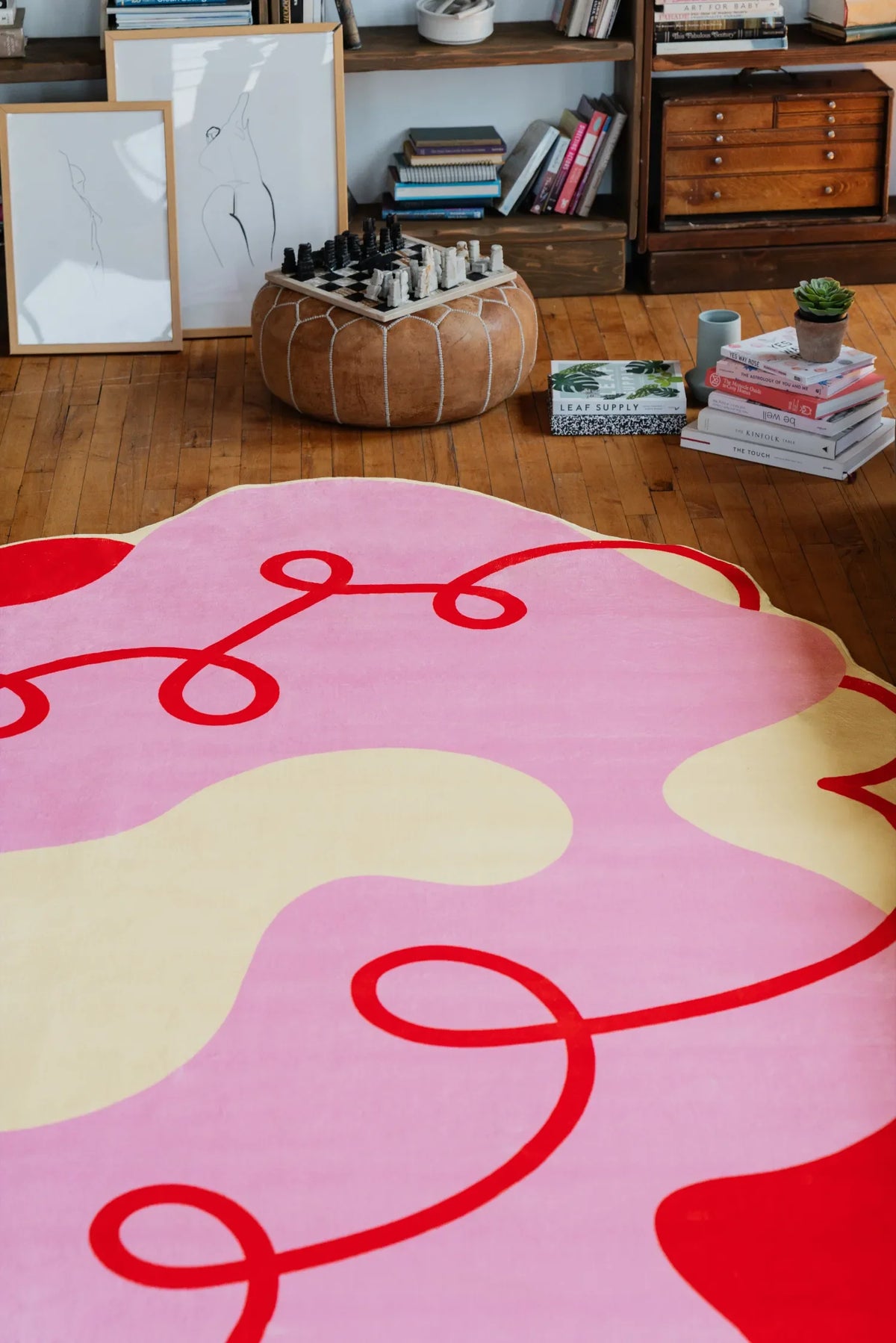 Pink funky area rug with red swirls, styled near a pouf, stacked books and framed art in a warm living room setting.