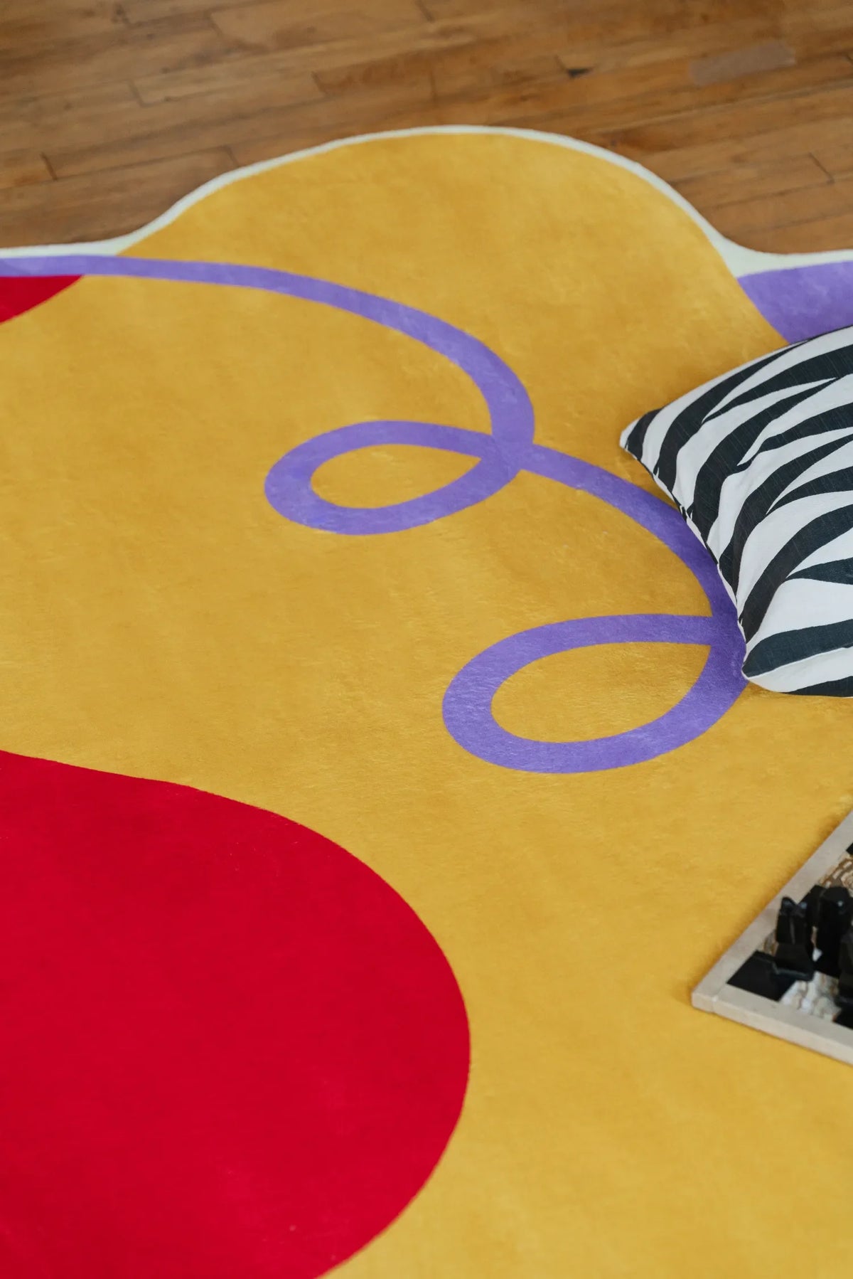 Close-up of the yellow abstract rug with purple swirls, set next to striped cushions and books on the wood floor.