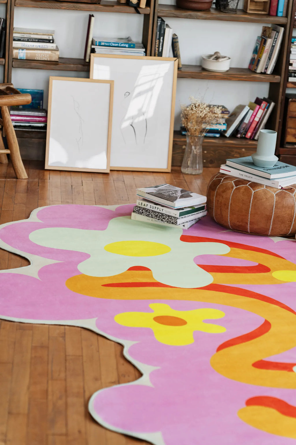 A funky floral rug in pink and orange tones on wood floor beside shelves and artwork leaning casually against the wall.