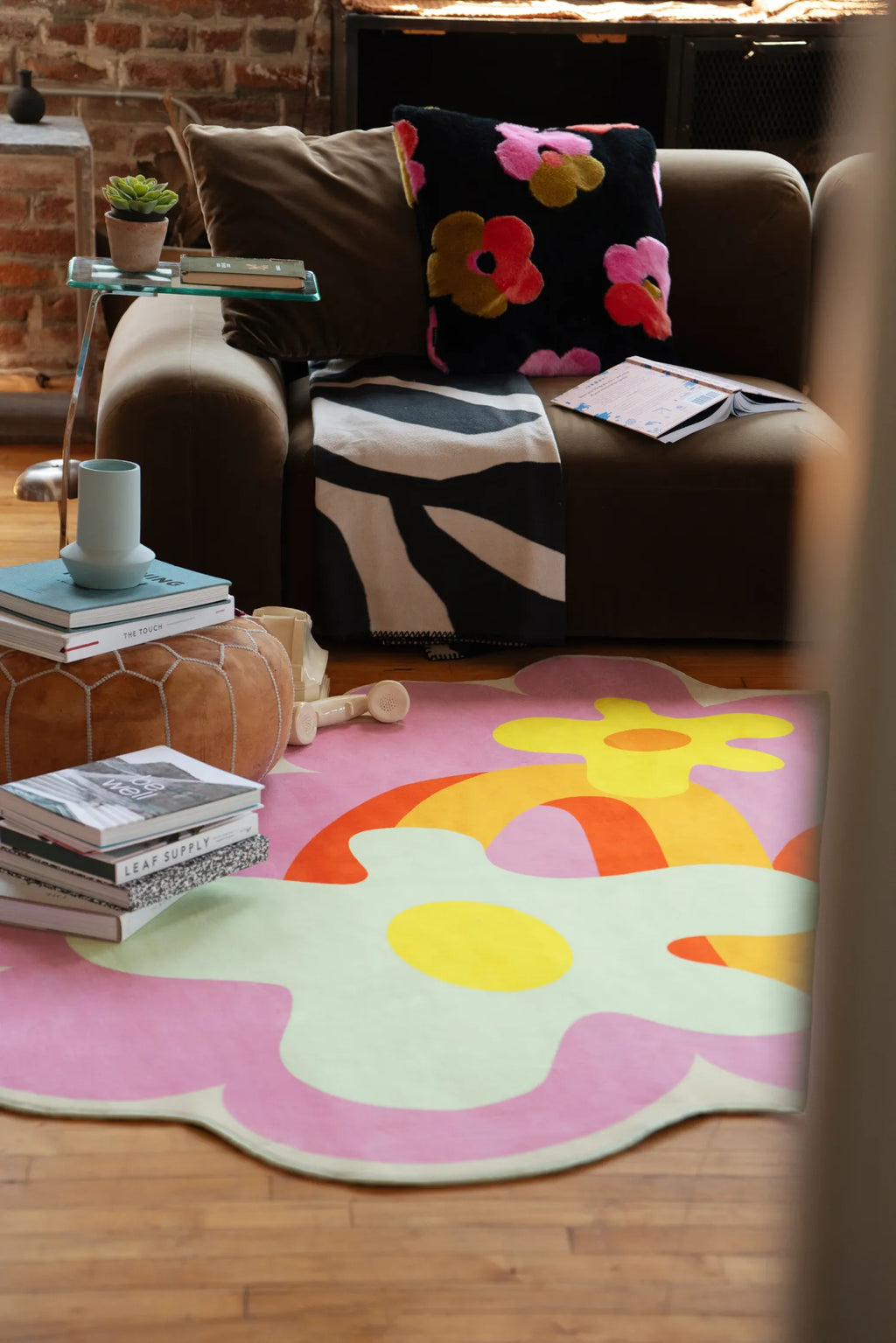 Pink floral rug with wavy design resting on wood floor in front of a dark sofa against exposed brick wall.